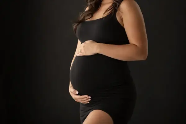Baby bump photoshoot 3 Maternity detail photography at PM STUDIOS Munich: Close-up of an expectant mother's baby bump as she gently cradles it with her hands, wearing a black dress against a dark studio background.