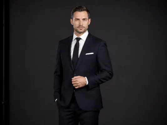 Businessfotos 3 Professional full-body business portrait by PM STUDIOS Munich: A man in a tailored dark navy suit with a black tie and white pocket square, standing against a dark studio background.