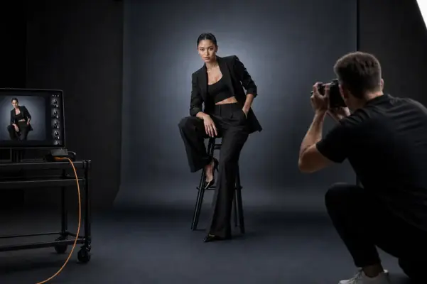 Professionelles Fashion-Shooting bei PM STUDIOS München: Ein Fotograf fotografiert ein Model im eleganten schwarzen Hosenanzug mit High Heels; Live-Bildkontrolle am Monitor und High-End Studiobeleuchtung.