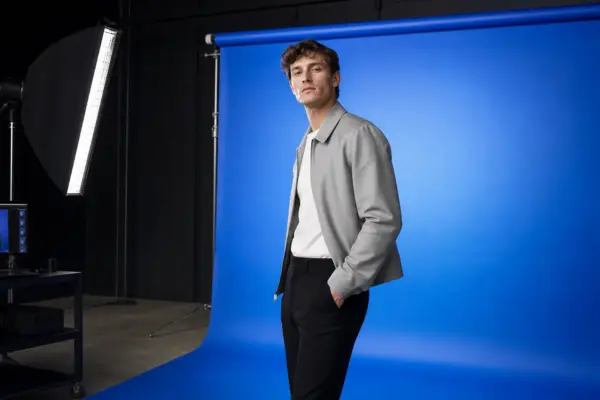 Professionelles Katalog-Shooting für Herrenmode bei PM STUDIOS München: Männliches Modell in grauer Jacke posiert vor einem kräftigen blauen Studiohintergrund; im Anschnitt professionelle Studiobeleuchtung und digitale Bildkontrollstation.