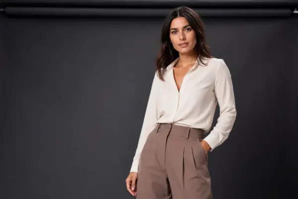 Comp card shooting 4 Professional studio portrait for a model comp card by PM STUDIOS Munich: Woman in an elegant cream blouse and brown trousers posing confidently against a dark grey background.