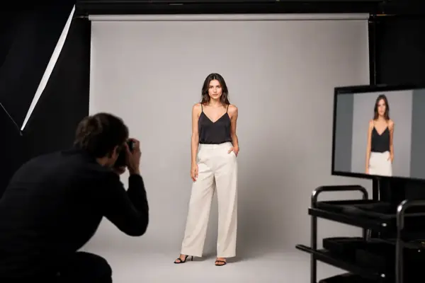 E-Commerce Shooting 1 Behind the scenes of an e-commerce fashion session at PM STUDIOS Munich: A photographer capturing a model in a black top and white trousers against a grey backdrop with live digital review on a studio monitor.