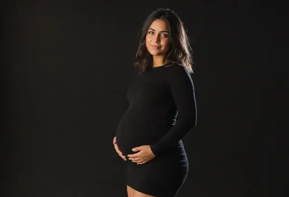 Baby bump photoshoot 1 Maternity portrait by PM STUDIOS Munich: A beautiful expectant mother in a form-fitting black dress posing against a dark studio background with elegant side lighting highlighting her baby bump.